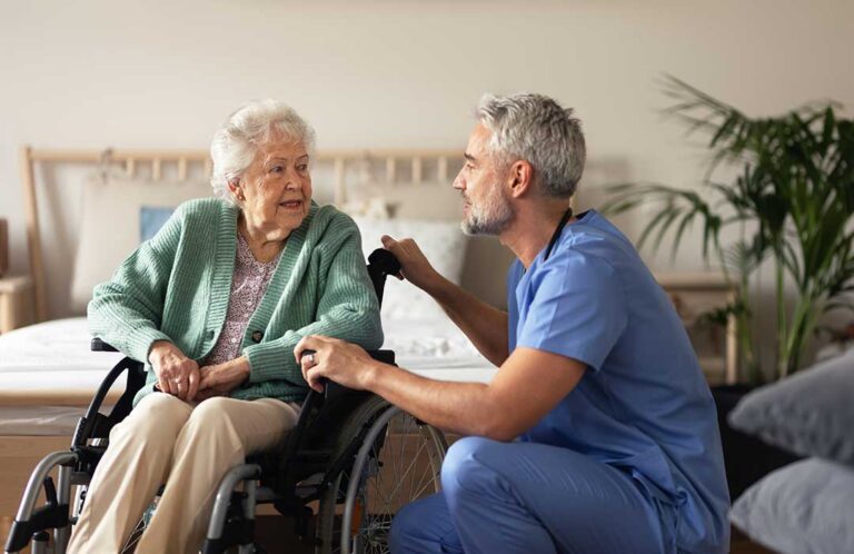 A group of older adults talking together in a light-filled common room, with personal belongings visible in the background.